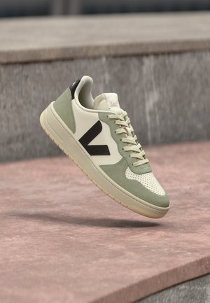 Light beige sneaker with green suede accents, perforated toe, and a dark brown logo. Rubber sole with textured grip. Lace-up design.
