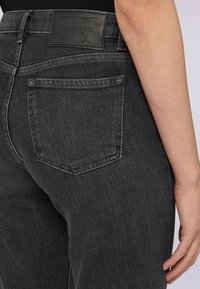 Vaqueros de mezclilla negra con una cintura de altura media, detalles en los bolsillos traseros y una etiqueta de cuero con "TRUSSARDI" en relieve. Textura suave, ligeramente desgastados.