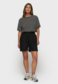 Mujer de pie vistiendo una camiseta a rayas blanca y negra, pantalones cortos negros y zapatillas plateadas frente a un fondo claro.