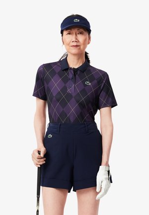 Paars en zwart argyle polo shirt met een kraagontwerp. Marineblauwe shorts met een subtiele vouw aan de zoom. Witte golfhandschoen en vizier. Golfclub aanwezig.