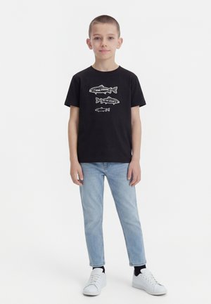 Αγόρι που στέκεται φορώντας μαύρο μπλουζάκι με τρία σχέδια ψαριών και φράσεις, ανοιχτό μπλε τζιν, μαύρες κάλτσες και λευκά αθλητικά παπούτσια.