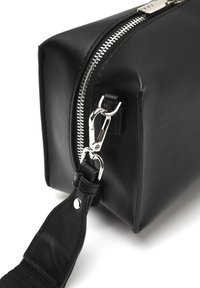 Sac en cuir noir avec fermeture à glissière argentée, de forme rectangulaire et avec une bandoulière amovible. Présente une texture brillante et des accents en chrome.