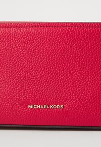 Piros bőr pénztárca texturált felülettel, arany "Michael Kors" logóval. Az élek feketérek, és a varrás látható a tetején.