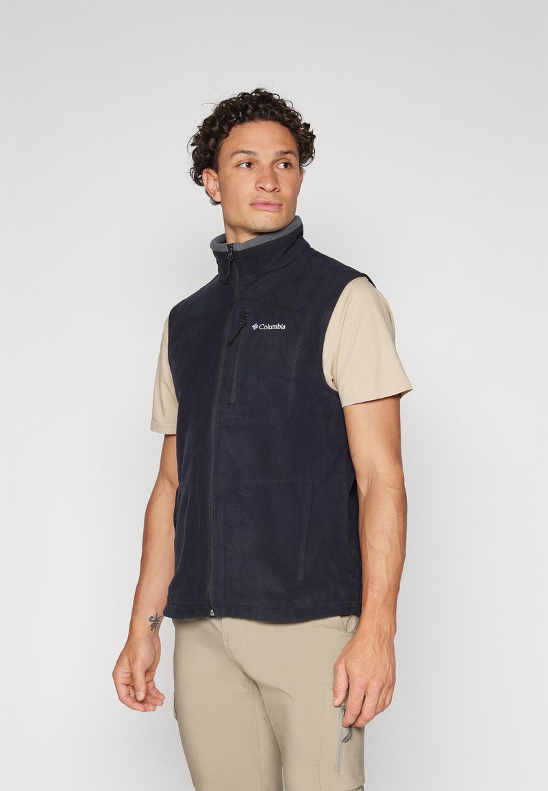 Navy fleece vest with high collar, zippered front, and two side pockets. Features subtle branding on the chest. Worn over a light beige shirt.