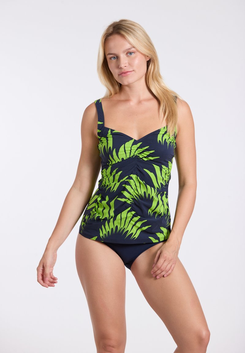 Femme blonde portant un haut de tankini bleu marine avec des motifs de feuilles tropicales vert vif et un bas de bikini bleu marine uni assorti.