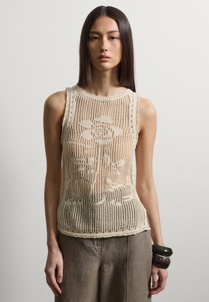 Frau trägt ärmelloses beiges Häkeloberteil mit Rosenmuster, braune Hose und große Armreifen am Handgelenk vor einfachem Hintergrund.