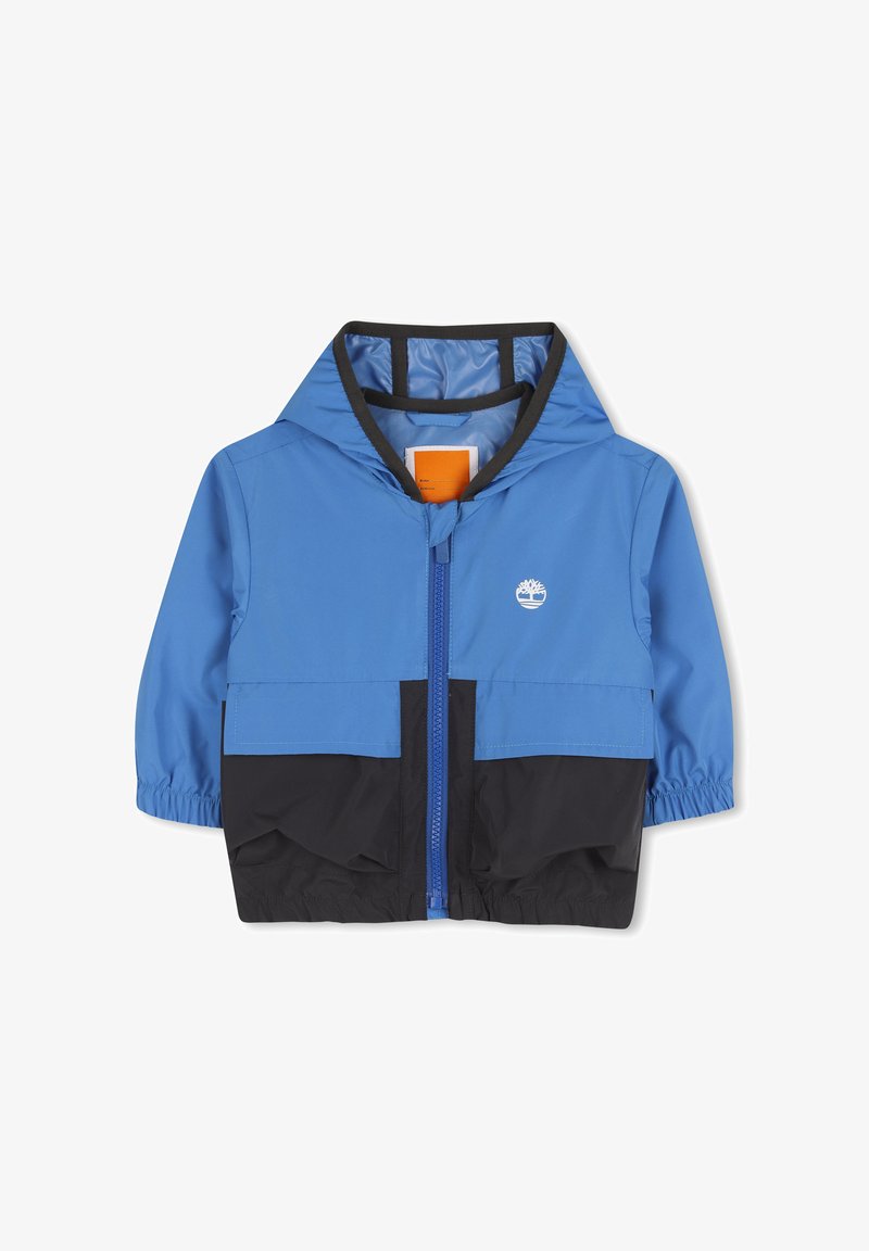 Veste coupe-vent bleue et noire avec capuche, fermeture éclair avant, poignets élastiques et logo sur la poitrine. Matière lisse et légère.