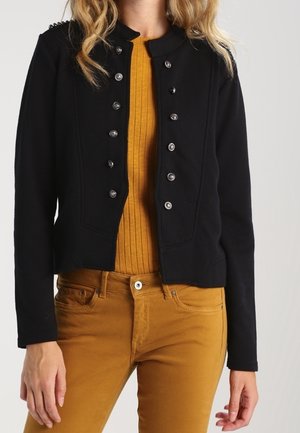 Veste noire courte avec un col rond et des boutons argentés. Tissu noir texturé, associée à un haut moutarde côtelé et à un pantalon.