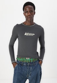 Γκρι μακρυμάνικο T-shirt με αντίθετο στρογγυλό λαιμό και λευκή γραφική εκτύπωση. Φοριέται με μπλε τζιν παντελόνι και πράσινη καρό ζώνη.