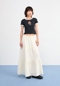 T camisa cropped preta com bordado de chamas e rosas, combinada com uma saia longa branca em camadas, com um padrão de grelha, e sapatos pretos.