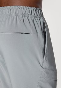Svetlo sivi športni hlače z rebrasto teksturo, z elastičnim pasom in žepi z zadrgo, označeni z "DRI-FIT" na pasu.