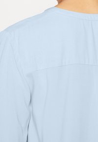 Chemise bleu clair à manches courtes avec une texture lisse, dotée d'une couture au dos et d'un léger drapé sur le tissu.