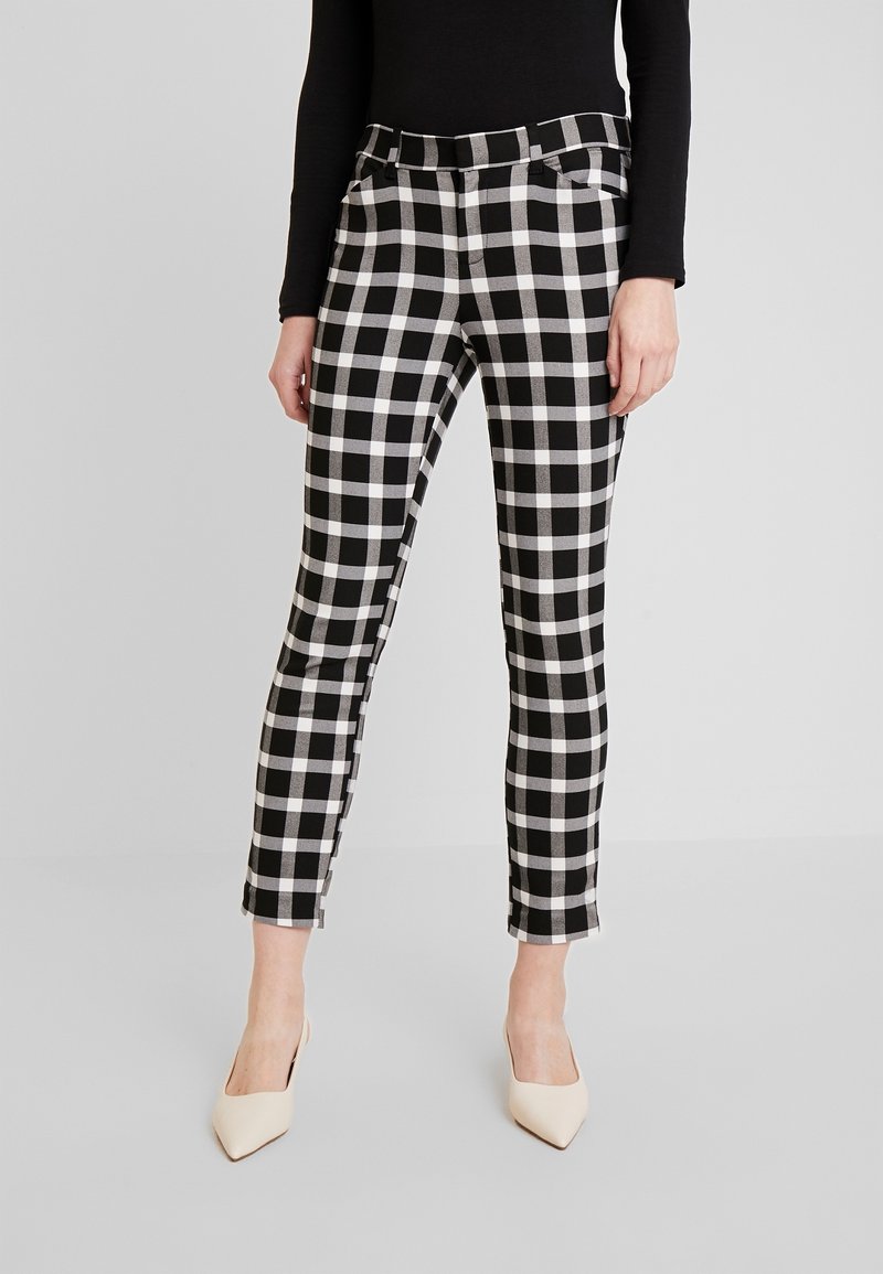 Femme portant un pantalon slim à carreaux noir et blanc avec un haut noir à manches longues et des talons pointus beiges.
