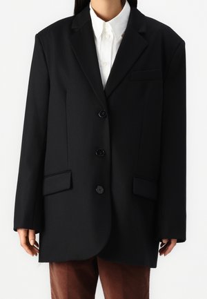 Osoba nosząca czarną oversize marynarkę na trzy guziki na białej koszuli z kołnierzykiem i brązowych spodniach, stojąca na jednolitym tle.