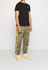 Person in einem schwarzen kurzärmeligen Shirt, olivgrünen Jogginghosen mit orangefarbenen Streifen an den Seiten, weißen Socken und beigen Sneakers.