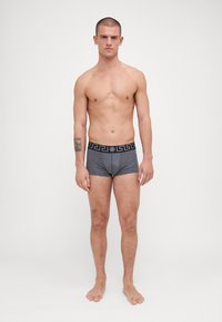 Grå boksershorts med svart midjebånd som har et gresk nøkkel mønster, laget av mykt stoff. Modell står barføtt med en nøytral bakgrunn.
