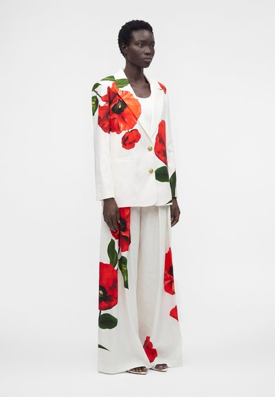 Femme portant un costume blanc avec de grandes fleurs de coquelicot rouges imprimées sur le blazer et un pantalon à jambes larges, debout devant un fond blanc.