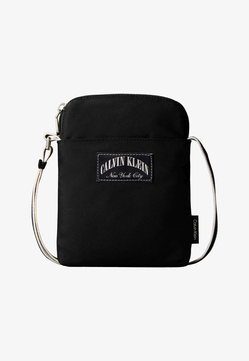 Μαύρη τσάντα ώμου Calvin Klein με λευκό ρυθμιζόμενο λουρί και μπροστινή ετικέτα με την επιγραφή "Calvin Klein New York City."