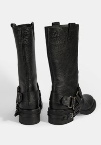 Zwarte leren knieholte laarzen met een gestructureerde afwerking, gespaccenten en een lage, stevige rubberen zool. Beschikt over treklabel voor gemakkelijke aantrekking.