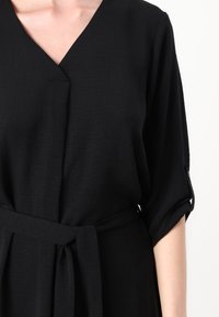 Schwarzes Kleid mit V-Ausschnitt, Dreiviertelärmeln und gebundenem Bund. Der Stoff hat eine strukturierte Oberfläche, die ein subtile Muster bietet.