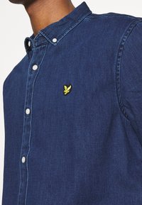 Camicia di jeans di colore blu scuro con colletto a punta, dotata di bottoni bianchi e un logo di uccello ricamato in giallo sul petto.