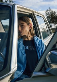 Sweat à capuche bleu clair zippé sur un t-shirt noir avec un texte blanc, assis dans une voiture, avec un cadre de porte métallique et la nature visible en arrière-plan.