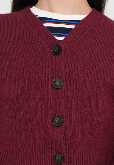 Burgundin värinen neulecardigan, jossa on V-pääntie, viisi mustaa nappia ja ribbireunus helmoissa ja hihansuissa. Raidallinen T-paita alla.