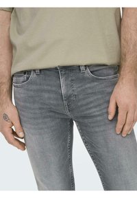 Jeans din denim gri cu o textură decolorată, cinci buzunare și fermoar. Modelul poartă un tricou de culoare verde măsliniu deschis și are un tatuaj pe mână.