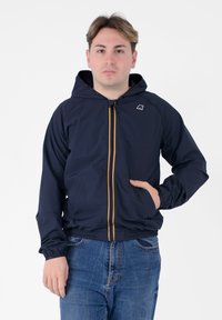 Giacca leggera navy con cappuccio, dotata di una cerniera multicolore, polsini elasticizzati e tasche frontali, indossata sopra jeans blu.