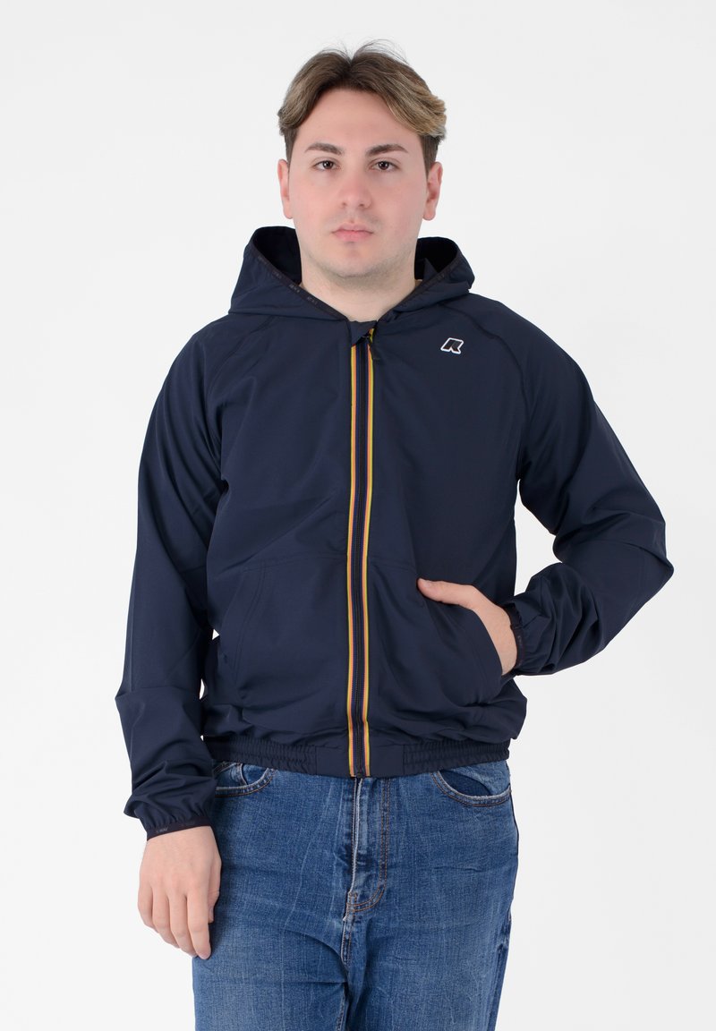 Giacca leggera navy con cappuccio, dotata di una cerniera multicolore, polsini elasticizzati e tasche frontali, indossata sopra jeans blu.