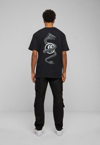 Črna bombažna majica s podrobno grafiko zmaja na hrbtu, v kombinaciji s črnimi cargo hlačami in belimi športnimi copati.