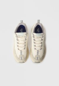 Gaiši bēšas sporta kurpes no auduma un zamša. Tās ir ar biezām šņorēm, pelēkām detaļām un teksturētu zolīti. Zīmola logotips redzams uz mēles.