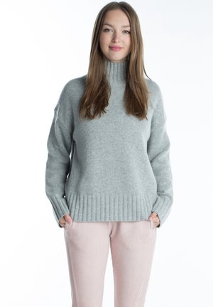 Jeune femme aux longs cheveux bruns portant un pull à col roulé gris clair et un pantalon rose clair, debout les mains dans les poches.