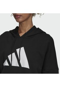 Černá mikina s kapucí, na které se nachází bílé logo se třemi pruhy, šňůrky na kapuci a volný střih. Materiál vypadá měkký a texturovaný.