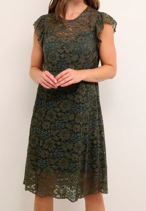 Mujer con vestido de encaje floral color verde oliva hasta la rodilla y mangas con volantes, de pie frente a un fondo claro y liso.