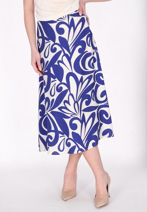 Femme portant une jupe blanche ornée de motifs floraux abstraits bleus audacieux et des chaussures à talons beiges, debout avec une main sur la hanche.