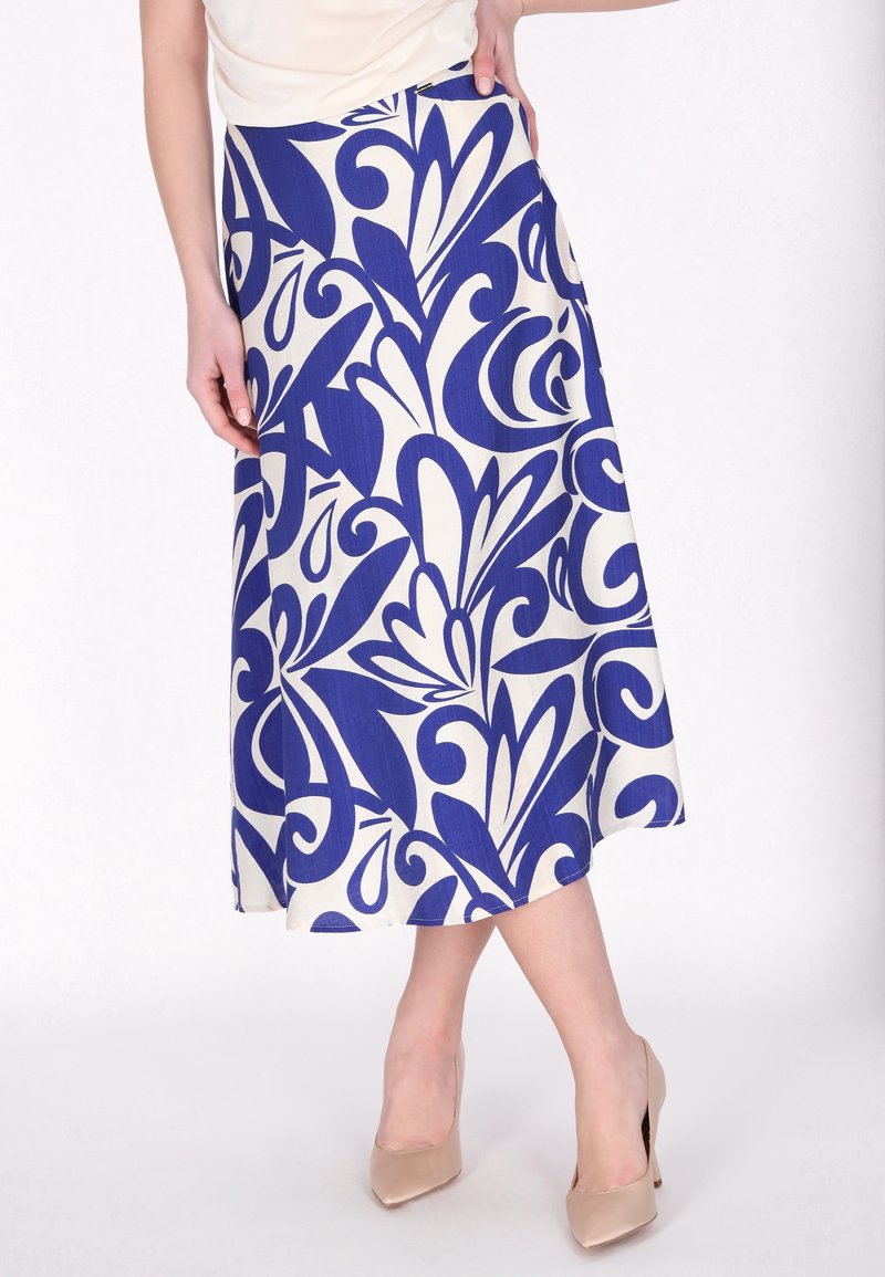 Femme portant une jupe blanche ornée de motifs floraux abstraits bleus audacieux et des chaussures à talons beiges, debout avec une main sur la hanche.