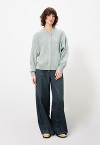 Hellgrüne, grob gestrickte Strickjacke mit V-Ausschnitt, kombiniert mit weit geschnittenen, dunkelgrauen Hosen und beigen Schuhen mit schwarzen Sohlen.