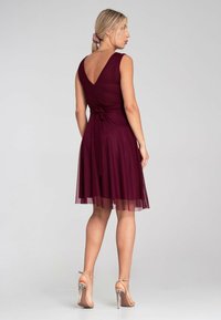 Burgunderfarbenes Kleid mit V-Ausschnitt, Taillengürtel und schichten Tüllrock. Durchsichtiger Saum mit fester Unterlage. Trägt transparente Absatzsandalen.