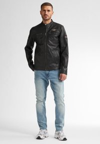Schwarze Lederjacke mit hohem Kragen, Reißverschluss und dekorativen Applikationen; kombiniert mit hellblauen Jeans und weißen Sportschuhen.