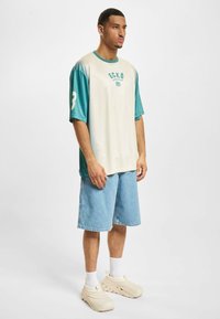 Romslig kortermet skjorte i krem og teal, med "ECKŌ"-logo og nummer 3. Kombinert med denimshorts og kremfargede sko.