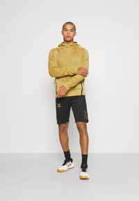 Gul huvtröja med svarta detaljer, tillsammans med svarta shorts med gula varumärkesloggor och chevronmönster, kompletterat med vita sneakers.