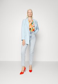 Blazer oversized azul claro sobre um top com padrão floral, jeans claros de cintura alta e sapatos de salto agudo vermelhos. Texturas suaves com cores vibrantes.