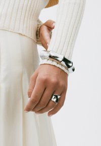 Hand med blänkande silverring och matchande kraftig armband som bärs över en krämfärgad ribbad tröja och kjol.