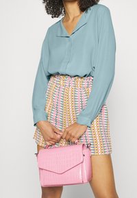 Femme portant un chemisier bleu clair et un short à motif multicolore, tenant un sac à main rose texturé avec les deux mains.