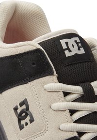 DC skate bateliai su smėlio spalvos zomšiniu viršumi, juodais megztiniais akcentais, baltais raišteliais ir ryškiu juodu logotipu šone.