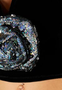 Musta samettivaate, jossa on kiiltävistä, monivärisistä paljeteista tehty spiraalimuotoilu ja hopeanvärinen korostus alareunassa.