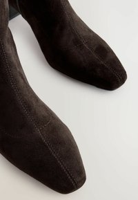 Botas de ante marrón hasta el tobillo con punta afilada y tacón de altura media. Presentan costuras visibles y una textura suave y lisa.
