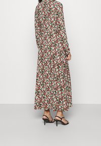 Donna che indossa un abito floreale a maniche lunghe, lungo fino alla caviglia, con fiori rossi, bianchi e verdi, abbinato a sandali neri con tacco alto e fasce.