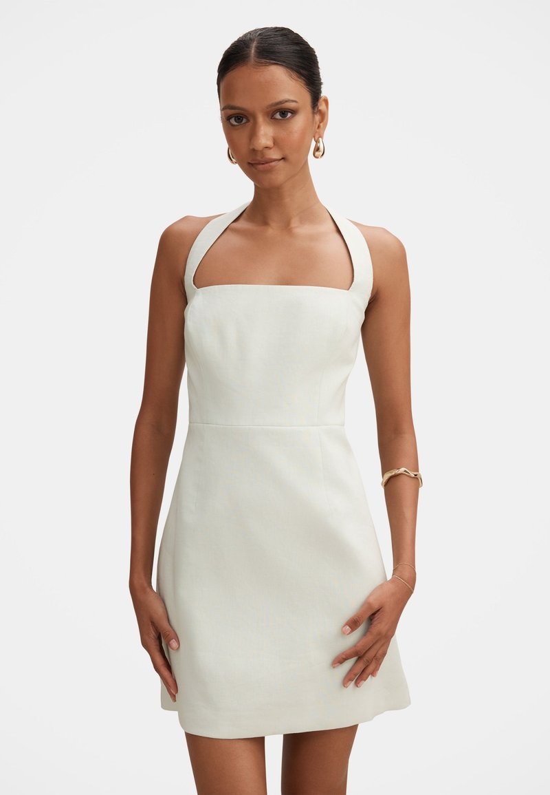 Γυναίκα με λευκό, αμάνικο φόρεμα με halter neck, χρυσά κρίκους ως σκουλαρίκια και δύο χρυσά βραχιόλια, στέκεται μπροστά σε απλό φόντο.